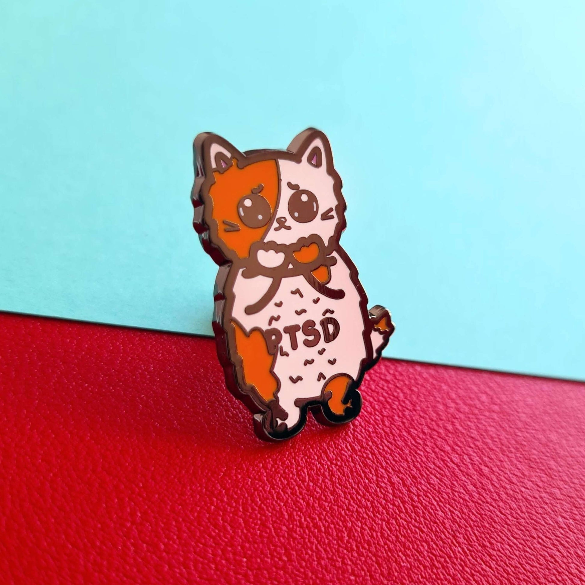 The PTSD Cat Enamel Pin - Post-Traumatic Stress Disorder on a red and blue background. An orange and white scared cat with black text across its belly reading 'PTSD'. The hand drawn design is raising awareness for post traumatic stress disorder.