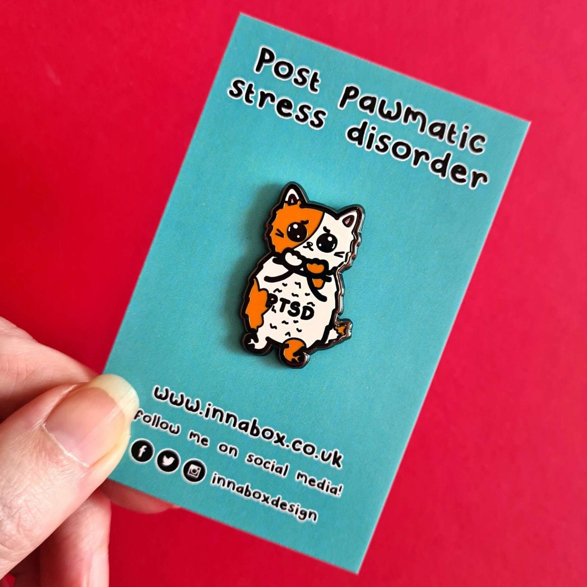 The PTSD Cat Enamel Pin - Post-Traumatic Stress Disorder on blue backing card with innabox social media handles underneath held over a red background. An orange and white scared cat with black text across its belly reading 'PTSD'. The hand drawn design is raising awareness for post traumatic stress disorder.