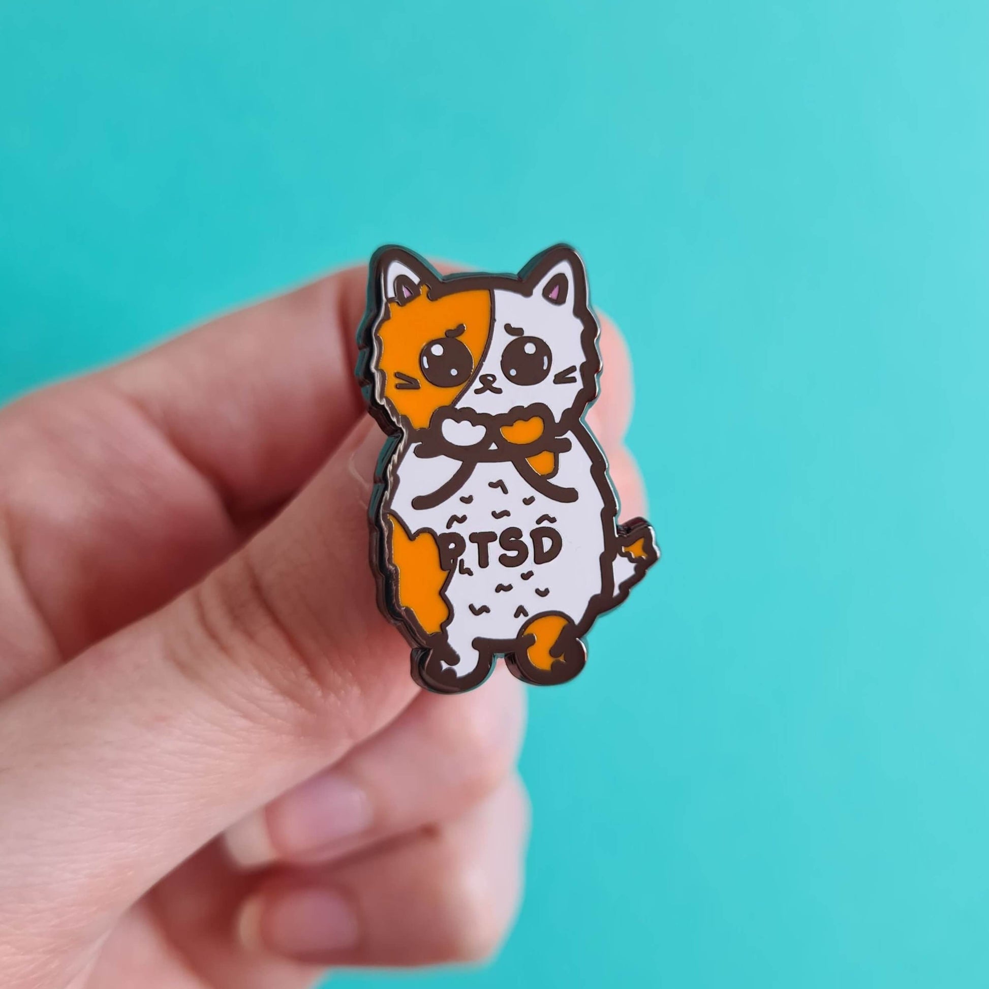 The PTSD Cat Enamel Pin - Post-Traumatic Stress Disorder held over a blue background. An orange and white scared cat with black text across its belly reading 'PTSD'. The hand drawn design is raising awareness for post traumatic stress disorder.