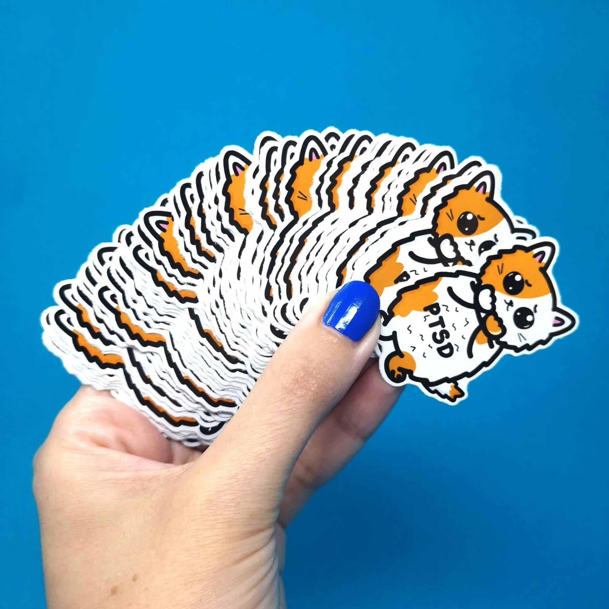 The PTSD Cat Sticker - Post-Traumatic Stress Disorder held in a pile over a blue background. The orange and white scared cat sticker has black text across its middle reading 'PTSD'. The hand drawn design is raising awareness for post traumatic stress disorder.