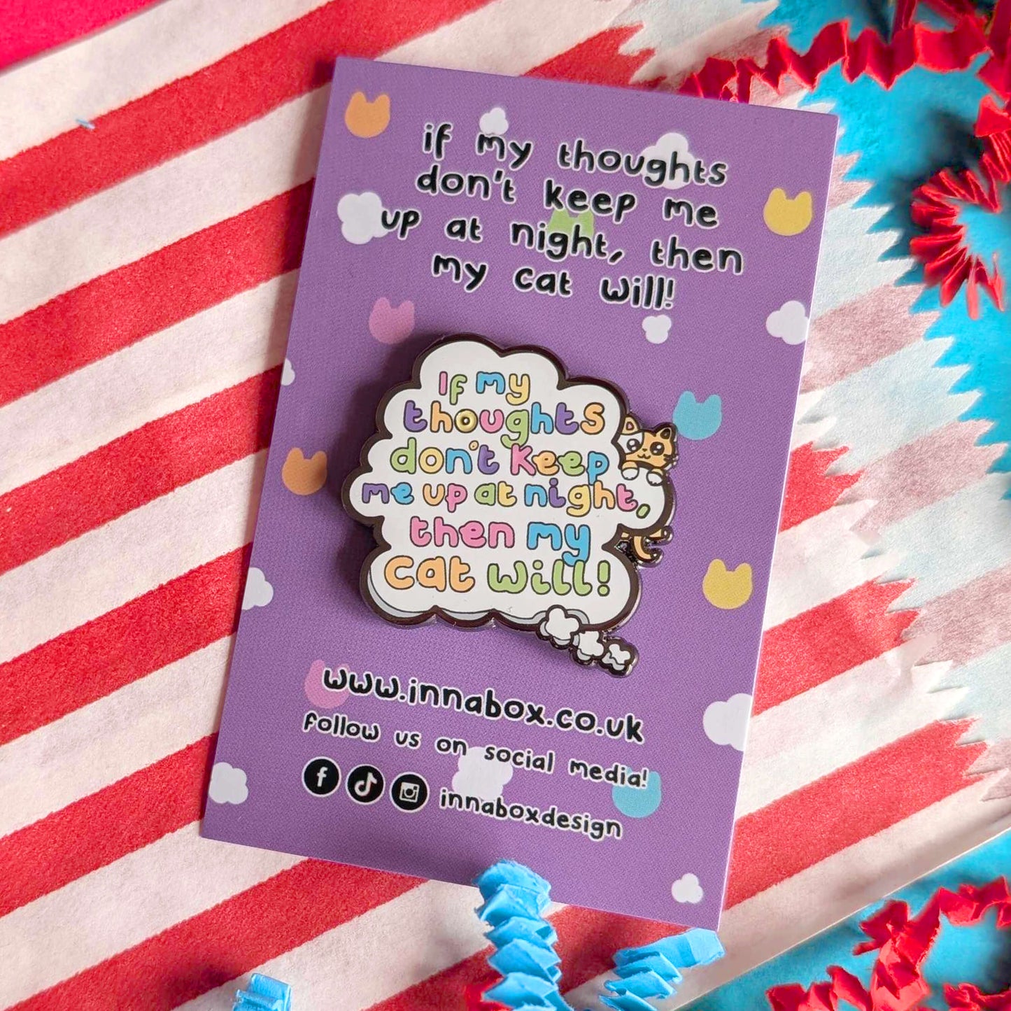 Innabox enamel pin featuring the phrase “If my thoughts don’t keep me up at night, then my cat will” on a lilac backing card, photographed on a red and white striped fabric with colourful paper accents. Playful cat themed pin supporting mental health awareness.
