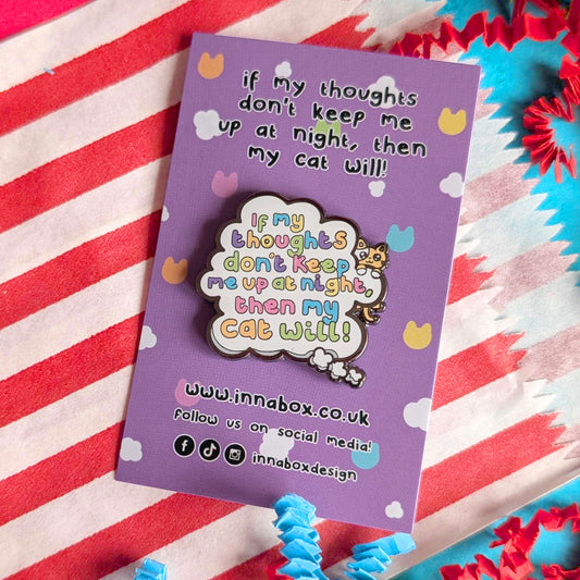Innabox enamel pin featuring the phrase “If my thoughts don’t keep me up at night, then my cat will” on a lilac backing card, photographed on a red and white striped fabric with colourful paper accents. Playful cat themed pin supporting mental health awareness.
