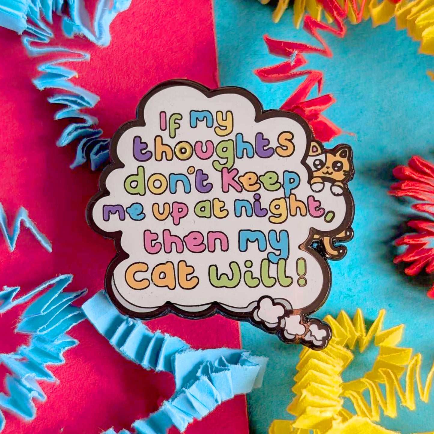 Close-up of Innabox enamel pin with multicoloured lettering reading “If my thoughts don’t keep me up at night, then my cat will”, featuring a cute illustrated cat and cloud design. Set against bold red and blue background with bright crinkle paper details.
