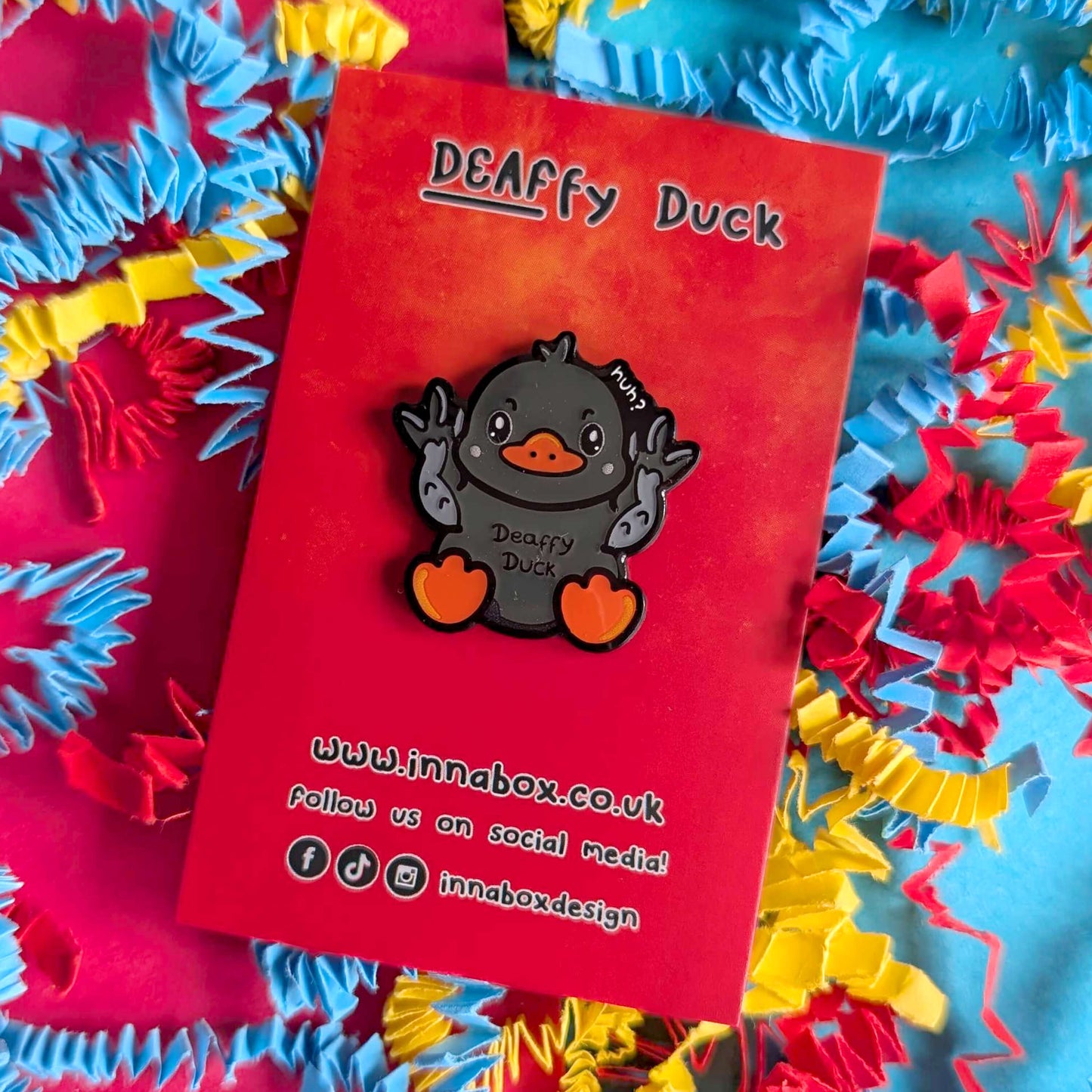 Deaffy Duck enamel pin by Innabox displayed on a vibrant red backing card with colourful crinkled paper background. The cute grey duck character signs BSL with raised hands, featuring orange beak and feet. Disability-positive enamel pin designed to celebrate Deaf culture and raise awareness of hidden disabilities.