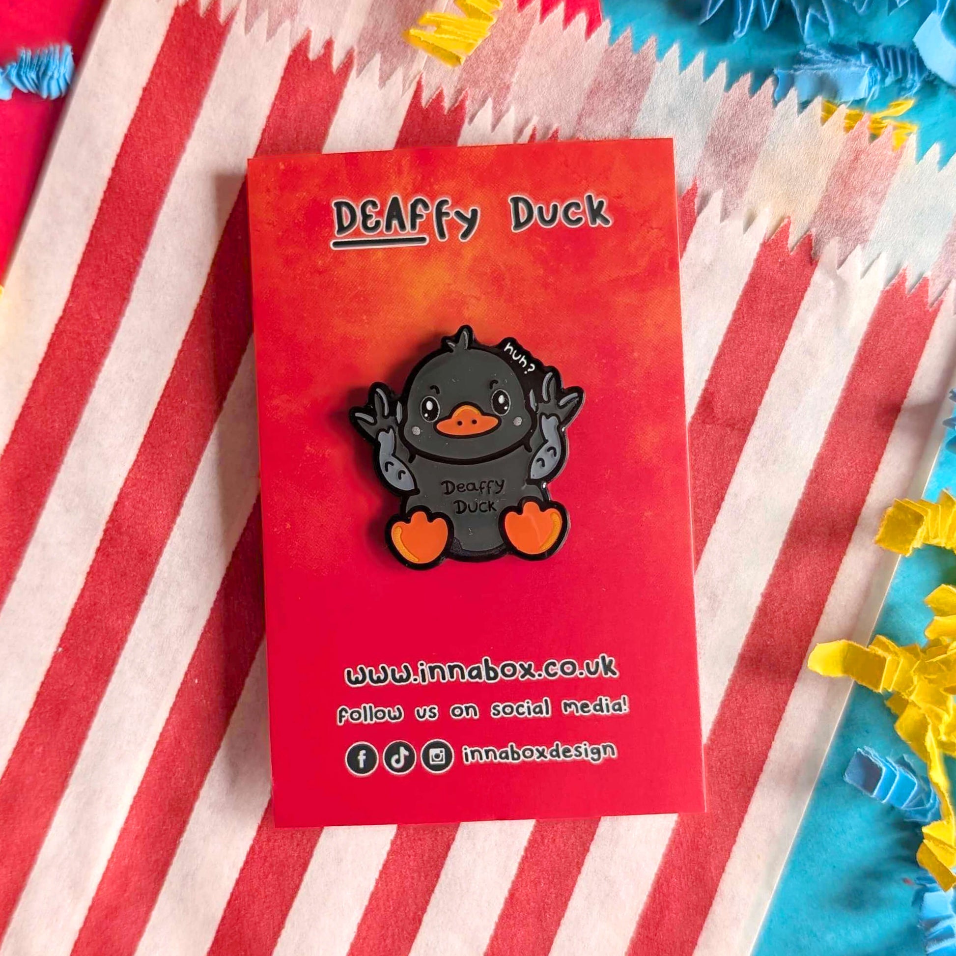 Deaffy Duck enamel pin by Innabox shown on a red branded backing card placed on red and white striped fabric. The grey duck character uses BSL hand gestures, with orange beak and feet. A fun, disability-inspired enamel pin promoting Deaf awareness and inclusive representation.
