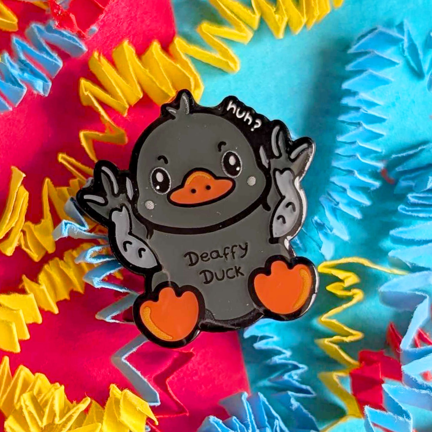 Close-up of Deaffy Duck enamel pin by Innabox against a colourful textured background. The grey duck character signs BSL with both hands raised, featuring orange beak and feet and a playful “huh?” detail. A cheerful enamel pin celebrating Deaf culture and disability awareness.
