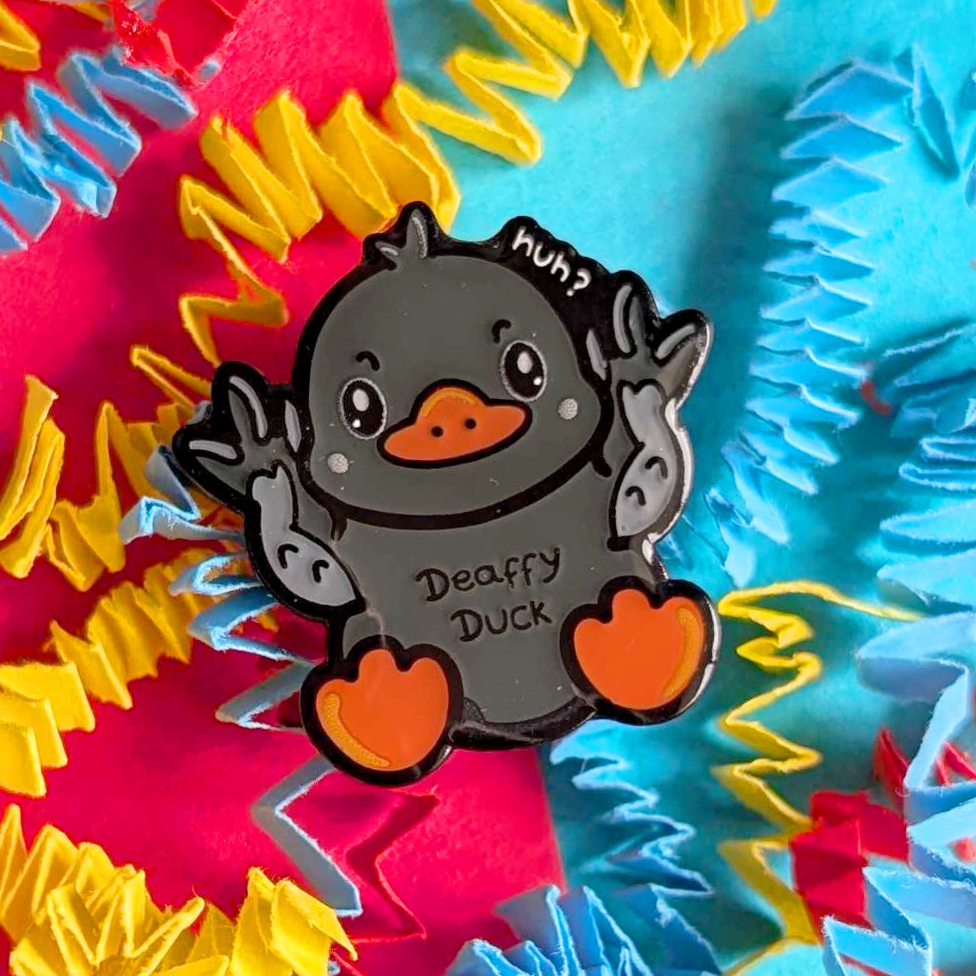 Close-up of Deaffy Duck enamel pin by Innabox against a colourful textured background. The grey duck character signs BSL with both hands raised, featuring orange beak and feet and a playful “huh?” detail. A cheerful enamel pin celebrating Deaf culture and disability awareness.
