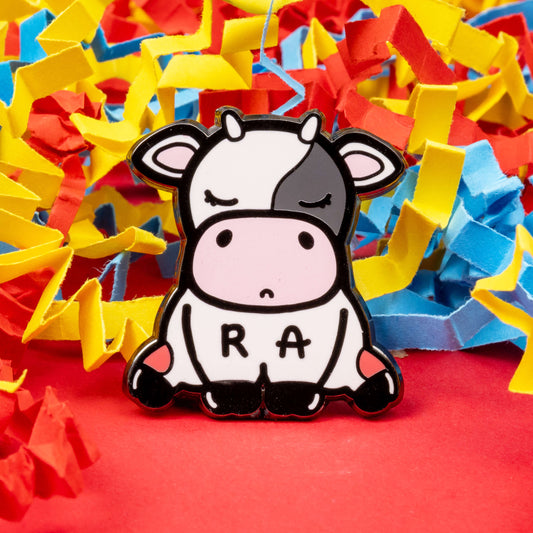 The Moomatoid Arthritis Cow Enamel Pin - Rheumatoid Arthritis on a red, yellow and blue card confetti background. The sad cow shape pin has red marks on its lower back legs and the initials R and A across its middle. The hand drawn design is raising awareness for Rheumatoid Arthritis.