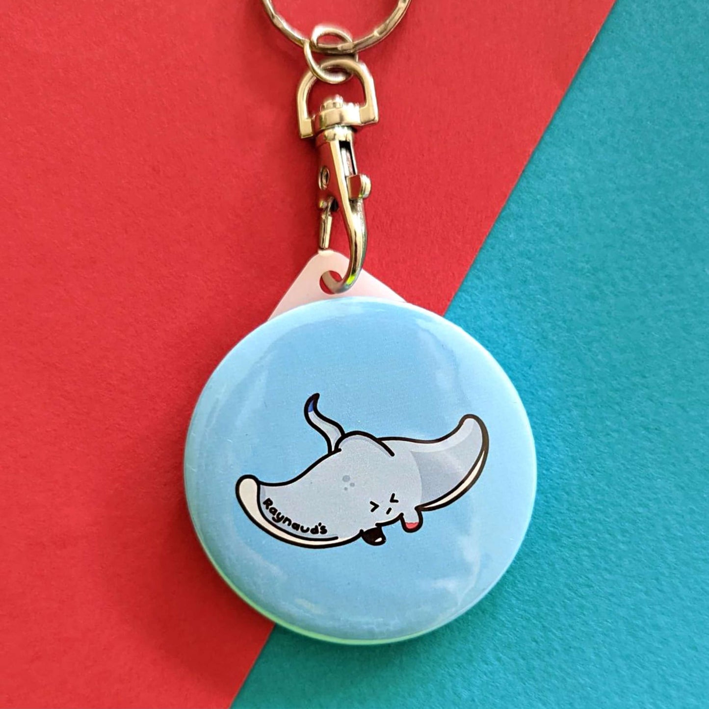 The Raynaud's Sting Ray Keyring on a red and blue background. The silver lobster clip blue plastic circular keychain with a grey sting ray with a scrunched sad face and red patches with black text across its wing reading 'raynaud's'. The hand drawn design is raising awareness for Raynaud's.
