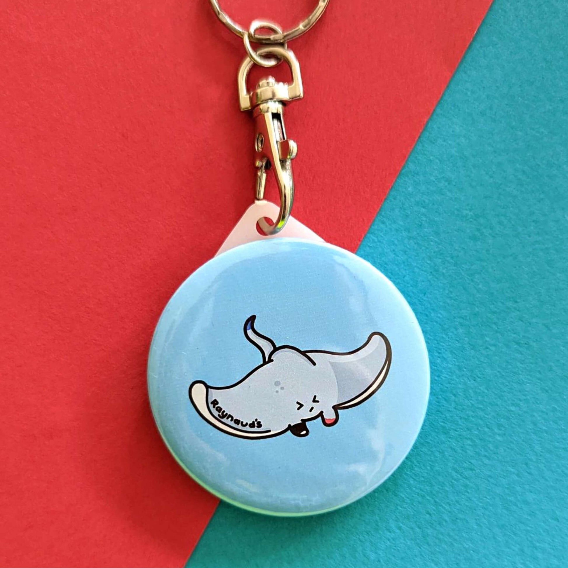 The Raynaud's Sting Ray Keyring on a red and blue background. The silver lobster clip blue plastic circular keychain with a grey sting ray with a scrunched sad face and red patches with black text across its wing reading 'raynaud's'. The hand drawn design is raising awareness for Raynaud's.