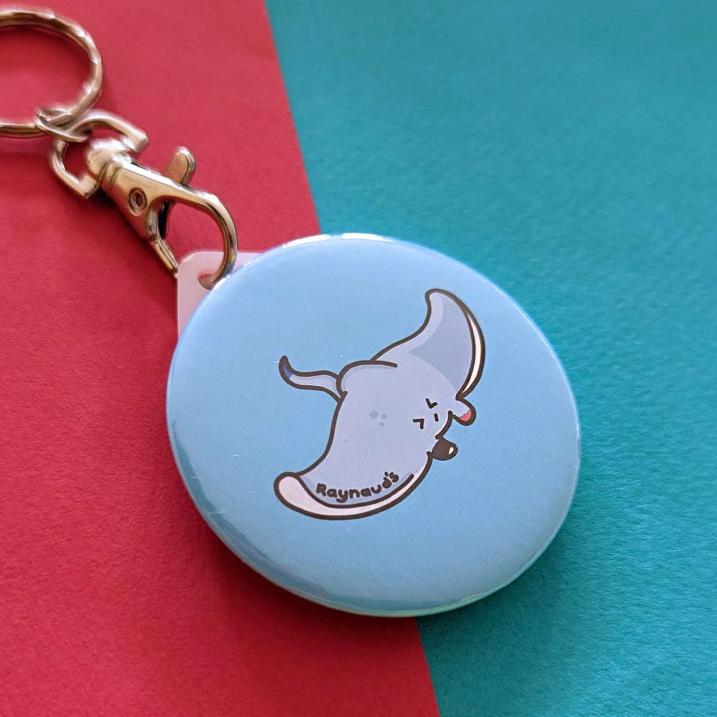 The Raynaud's Sting Ray Keyring on a red and blue background. The silver lobster clip blue plastic circular keychain with a grey sting ray with a scrunched sad face and red patches with black text across its wing reading 'raynaud's'. The hand drawn design is raising awareness for Raynaud's.
