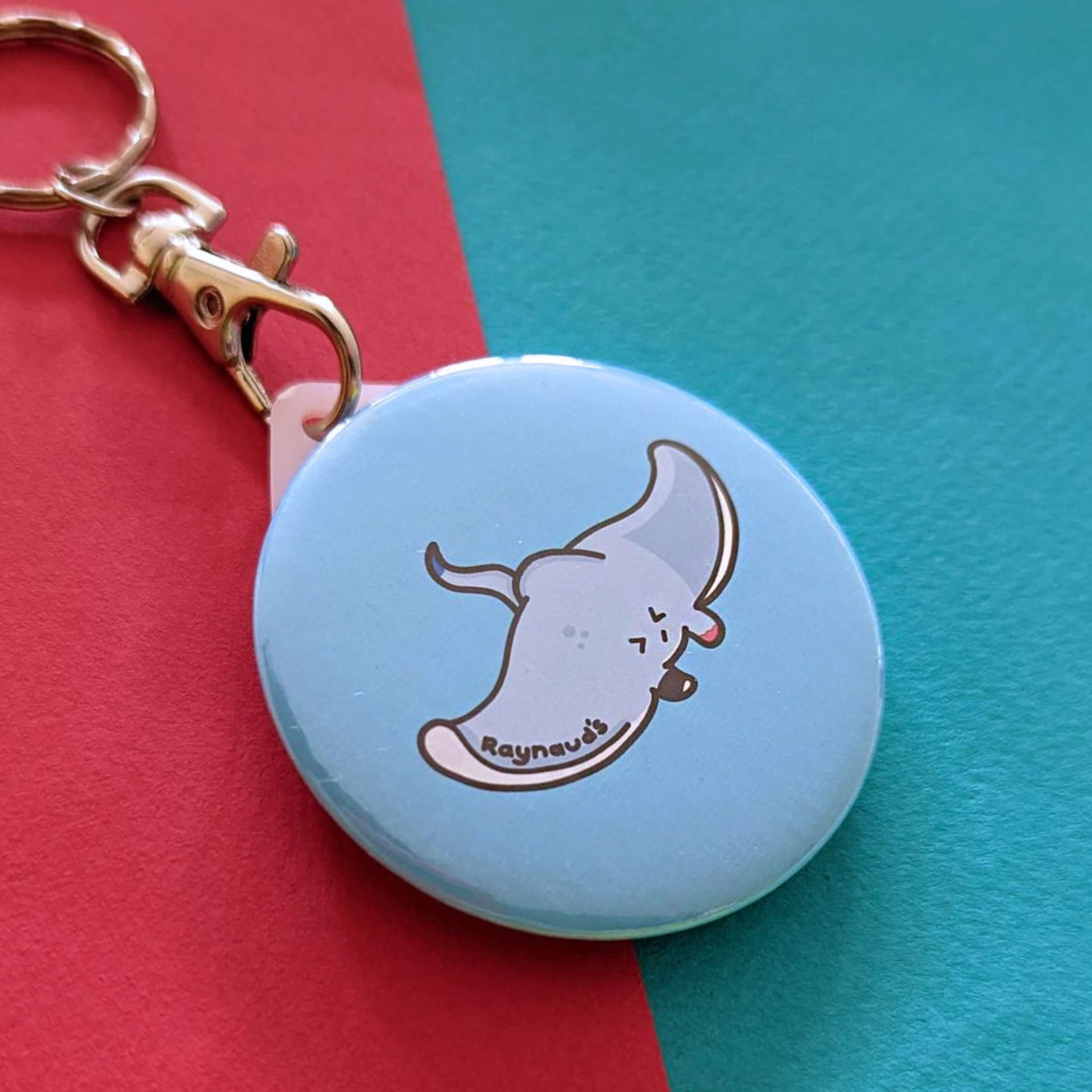 The Raynaud's Sting Ray Keyring on a red and blue background. The silver lobster clip blue plastic circular keychain with a grey sting ray with a scrunched sad face and red patches with black text across its wing reading 'raynaud's'. The hand drawn design is raising awareness for Raynaud's.