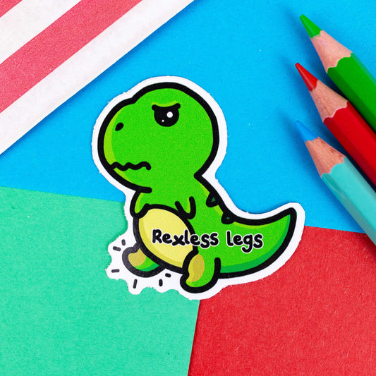 The Rexless Legs Sticker - Restless Legs Syndrome on a red, blue and green background with colouring pencils and red stripe candy bag. The green, grumpy upset t-rex dinosaur sticker has yellow lower legs with black text across its body reading 'rexless legs'. The hand drawn design is raising awareness for restless legs syndrome.