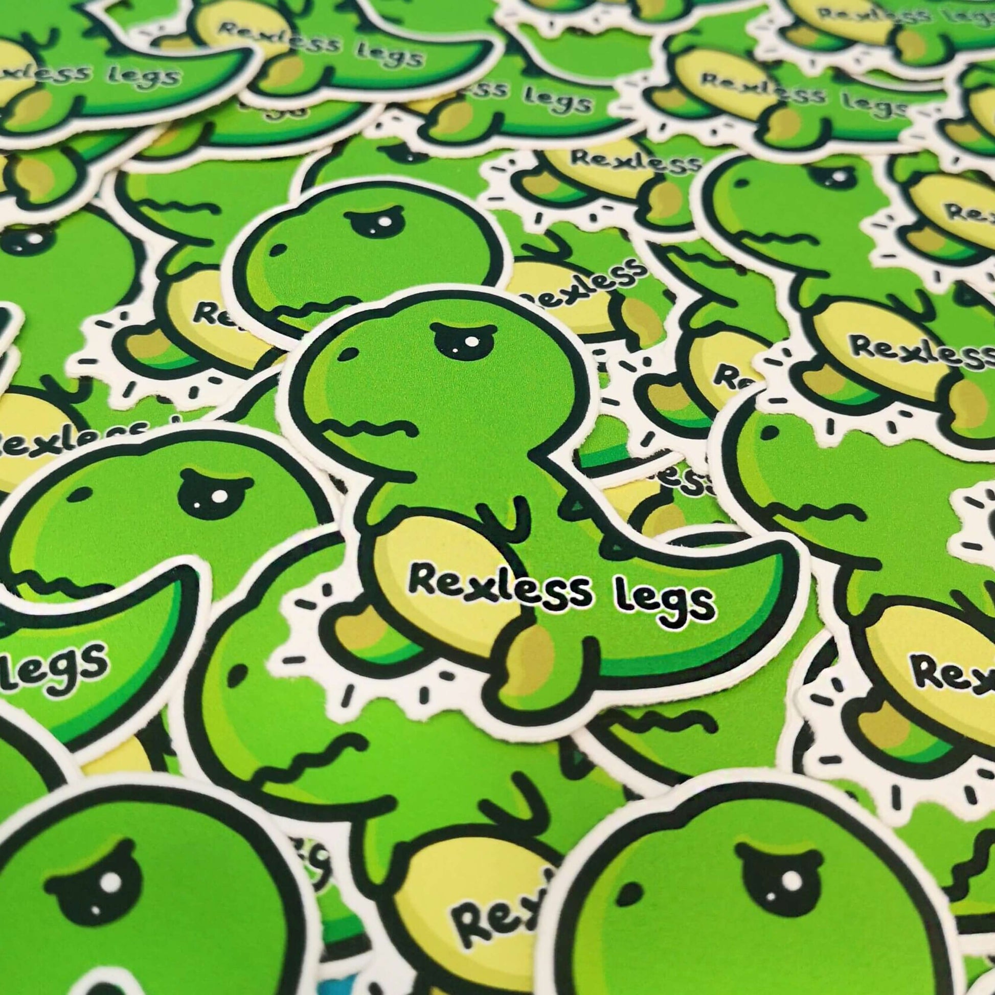 The Rexless Legs Sticker - Restless Legs Syndrome on a pile of the same copy of the sticker. The green, grumpy upset t-rex dinosaur sticker has yellow lower legs with black text across its body reading 'rexless legs'. The hand drawn design is raising awareness for restless legs syndrome.