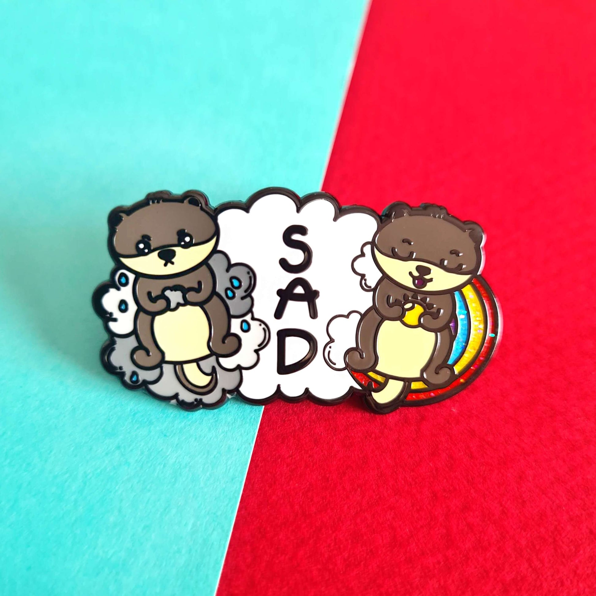 The Seasonal Affective Otter Enamel Pin - Seasonal Affective Disorder SAD on a red and blue background. The cloud shaped pin has two otters on either side, one sad on a raincloud clutching a raincloud and the other smiling on a rainbow clutching a sunshine. In the middle is the initials 'SAD'. The hand drawn design is raising awareness for Seasonal Affective Disorder.