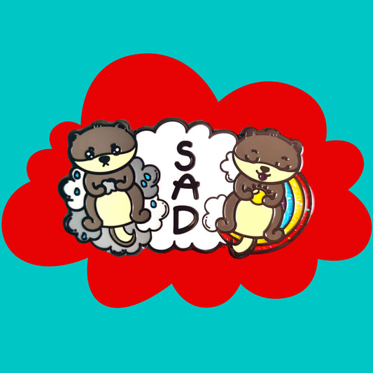 The Seasonal Affective Otter Enamel Pin - Seasonal Affective Disorder SAD on a red and blue background. The cloud shaped pin has two otters on either side, one sad on a raincloud clutching a raincloud and the other smiling on a rainbow clutching a sunshine. In the middle is the initials 'SAD'. The hand drawn design is raising awareness for Seasonal Affective Disorder.