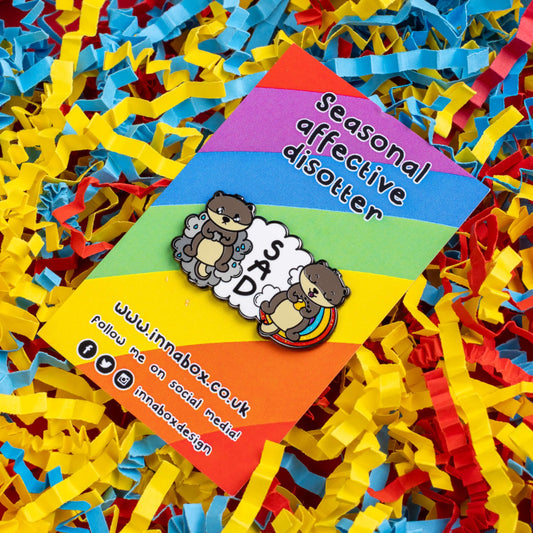 The Seasonal Affective Otter Enamel Pin - Seasonal Affective Disorder SAD on rainbow backing card laid on a red, yellow and blue card confetti background. The cloud shaped pin has two otters on either side, one sad on a raincloud clutching a raincloud and the other smiling on a rainbow clutching a sunshine. In the middle is the initials 'SAD'. The hand drawn design is raising awareness for Seasonal Affective Disorder.