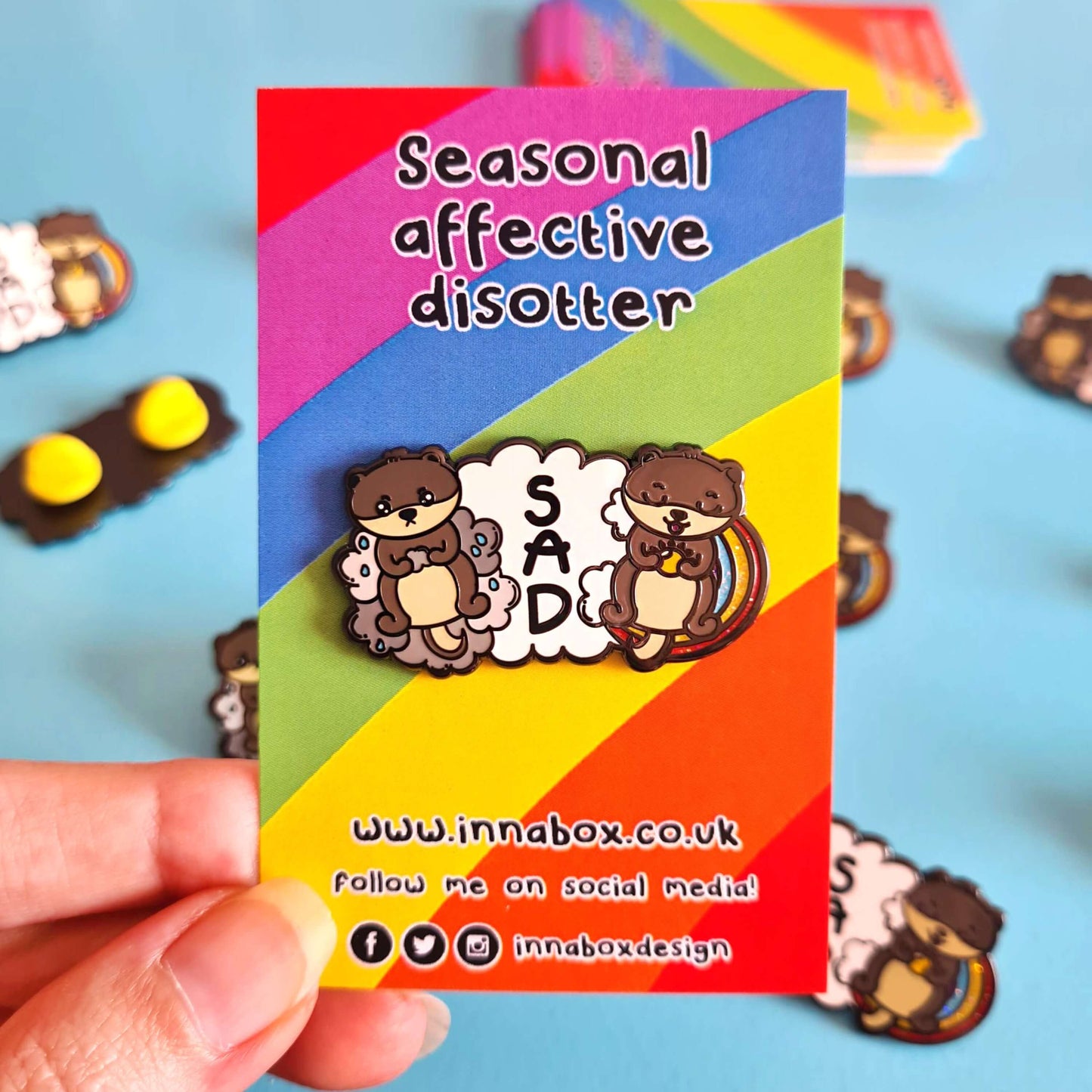 The Seasonal Affective Otter Enamel Pin - Seasonal Affective Disorder SAD on rainbow backing card being held over a blue background and multiple copies of the pin. The cloud shaped pin has two otters on either side, one sad on a raincloud clutching a raincloud and the other smiling on a rainbow clutching a sunshine. In the middle is the initials 'SAD'. The hand drawn design is raising awareness for Seasonal Affective Disorder.