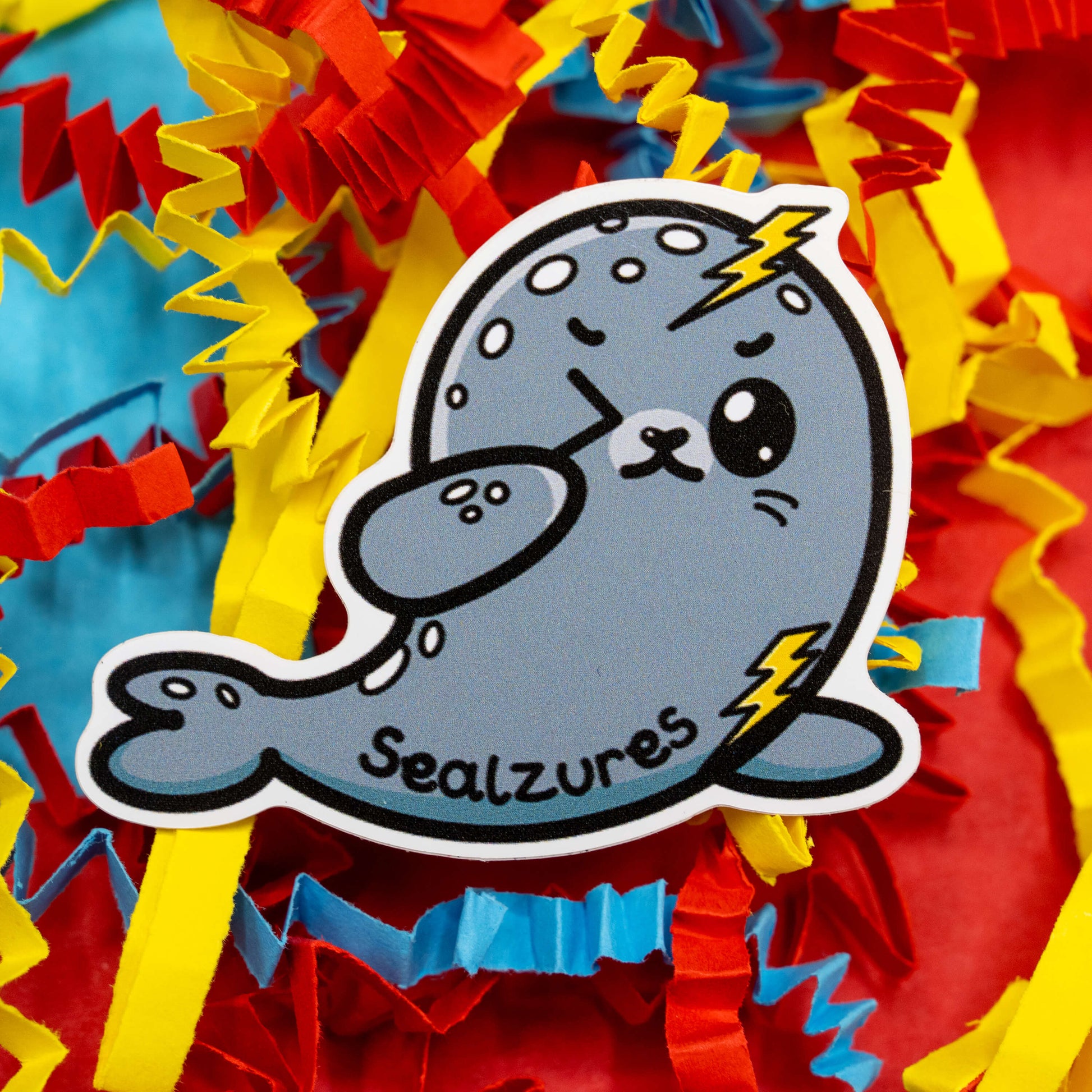 A cute grey seal cartoon character called Sealzures designed by Innabox. The seal has lightning bolts on its head and flipper, representing seizure awareness, and is surrounded by bright red, yellow, and blue confetti. This 5 cm gloss vinyl sticker celebrates hidden disabilities and chronic illness with a kind and humorous twist. Waterproof and suitable for indoor or outdoor use.