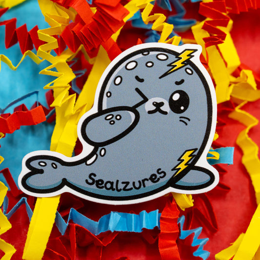 A cute grey seal cartoon character called Sealzures designed by Innabox. The seal has lightning bolts on its head and flipper, representing seizure awareness, and is surrounded by bright red, yellow, and blue confetti. This 5 cm gloss vinyl sticker celebrates hidden disabilities and chronic illness with a kind and humorous twist. Waterproof and suitable for indoor or outdoor use.