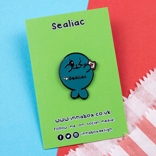 The Sealiac Seal Enamel Pin - Coeliac Disease on green backing card laying on a red and blue background. A blue winking smiling seal holding up a piece of toast with a red cross, across its belly in black text reads 'sealiac'. The hand drawn design is raising awareness for coeliac disease.