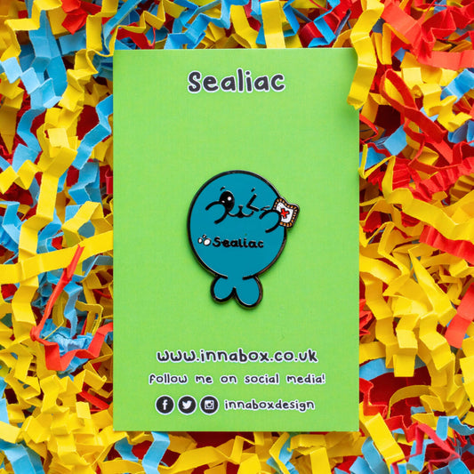 The Sealiac Seal Enamel Pin - Coeliac Disease on green backing card laying on a red, yellow and blue card confetti background. A blue winking smiling seal holding up a piece of toast with a red cross, across its belly in black text reads 'sealiac'. The hand drawn design is raising awareness for coeliac disease.