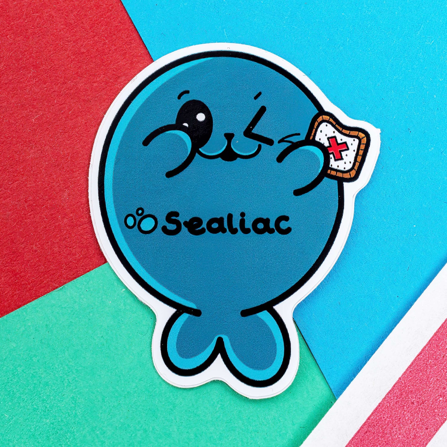 The Sealiac Seal Sticker - Coeliac Disease on a red, blue and green background with a red stripe candy bag. The winking blue seal sticker is holding up a piece of toast with a red cross, along its belly in black text reads 'sealiac'. The hand drawn design is raising awareness for Coeliac Disease.