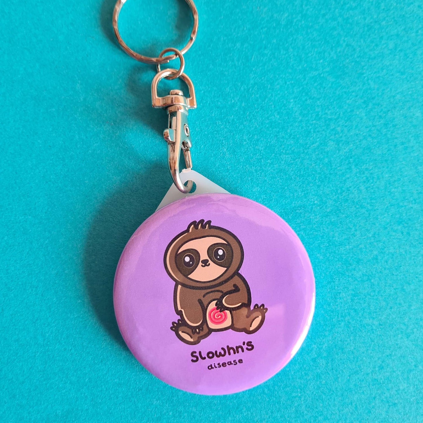 The Slowhn's Disease Sloth Keyring - Crohn's Disease on a blue background. The silver lobster clip pink plastic circular keyring of a smiling sat down brown sloth clutching its swirly red tummy and black text underneath reading 'slowhn's disease'. The hand made design is raising awareness for crohn's disease.