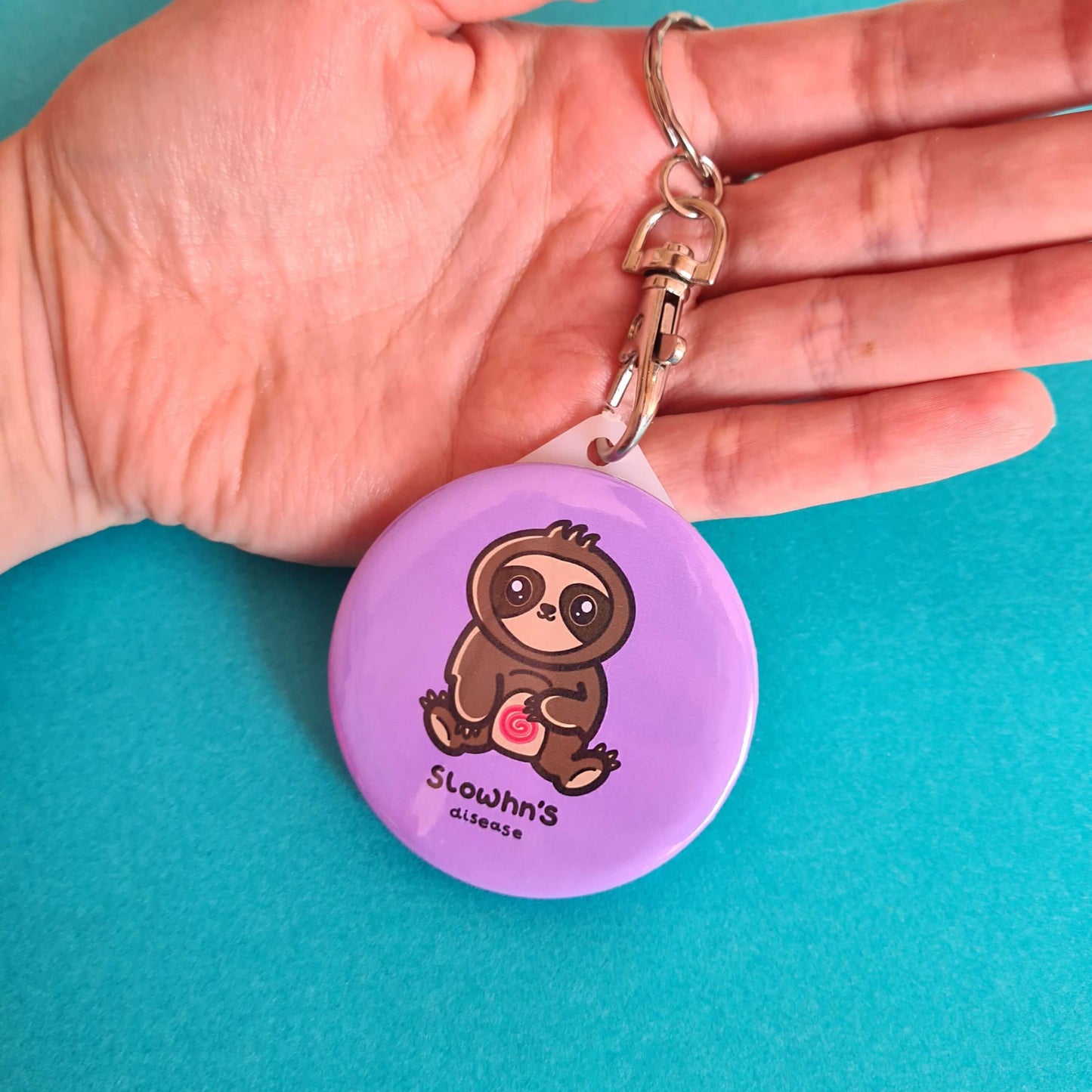 The Slowhn's Disease Sloth Keyring - Crohn's Disease held on a blue background. The silver lobster clip pink plastic circular keyring of a smiling sat down brown sloth clutching its swirly red tummy and black text underneath reading 'slowhn's disease'. The hand made design is raising awareness for crohn's disease.