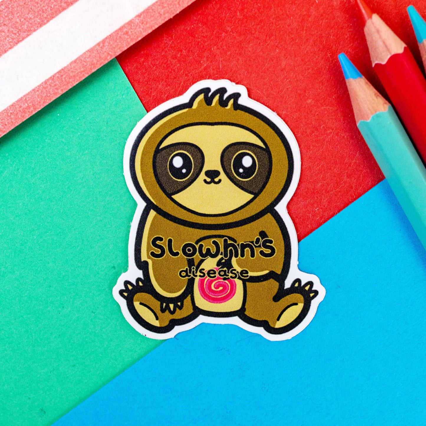The Slowhn's Disease Sloth Sticker - Crohn's Disease on a red, blue and green background with colouring pencils and a red stripe candy bag. The sloth shape sticker is smiling sat down clutching its tummy that has a red swirl and text reading 'slowhn's disease'. The design is raising awareness for crohn's disease.