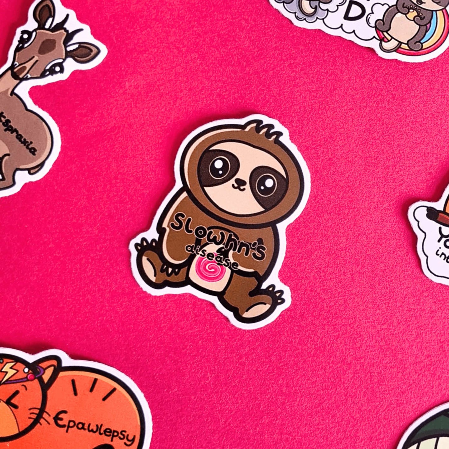 The Slowhn's Disease Sloth Sticker - Crohn's Disease being on a red background with other innabox stickers. The sloth shape sticker is smiling sat down clutching its tummy that has a red swirl and text reading 'slowhn's disease'. The design is raising awareness for crohn's disease.