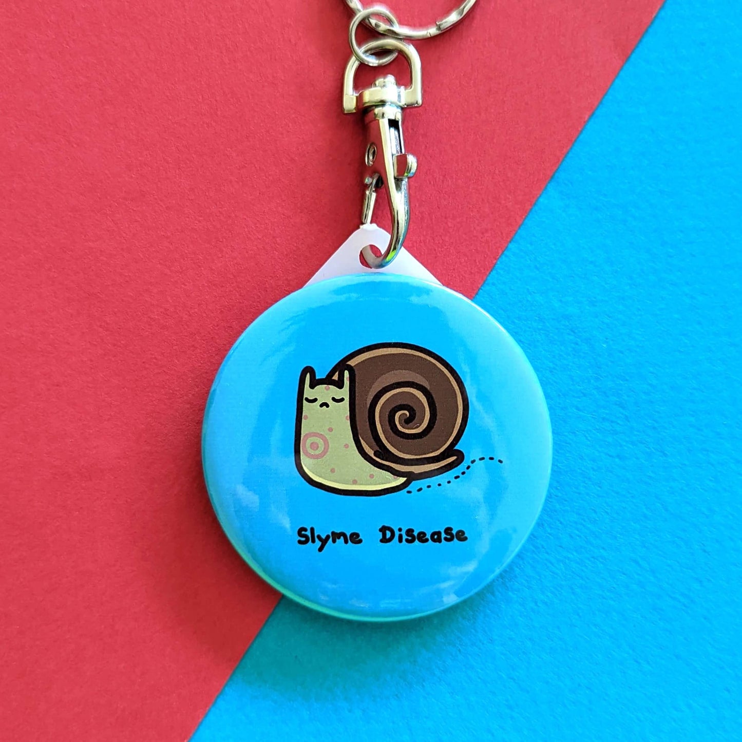 Slyme Disease Snail Keyring - Lyme Disease on a red and blue background. The pastel blue plastic circular keychain with silver lobster clip features a sad brown snail with red spots, underneath is black text reading 'slyme disease'. The hand drawn design is raising awareness for lyme disease.
