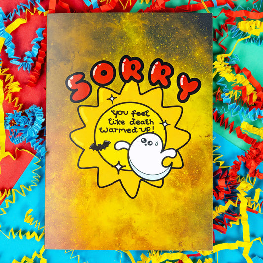 A yellow greeting card with black paint splatters. 'Sorry' is written in big red letters above an illustration of a sun with 'You feel like death warmed up!' inside. there are also sparkles, a sweating ghost and a bat around the sun. The background of the photo is colourful card confetti. The spooky design is inspired by get well soon cards.