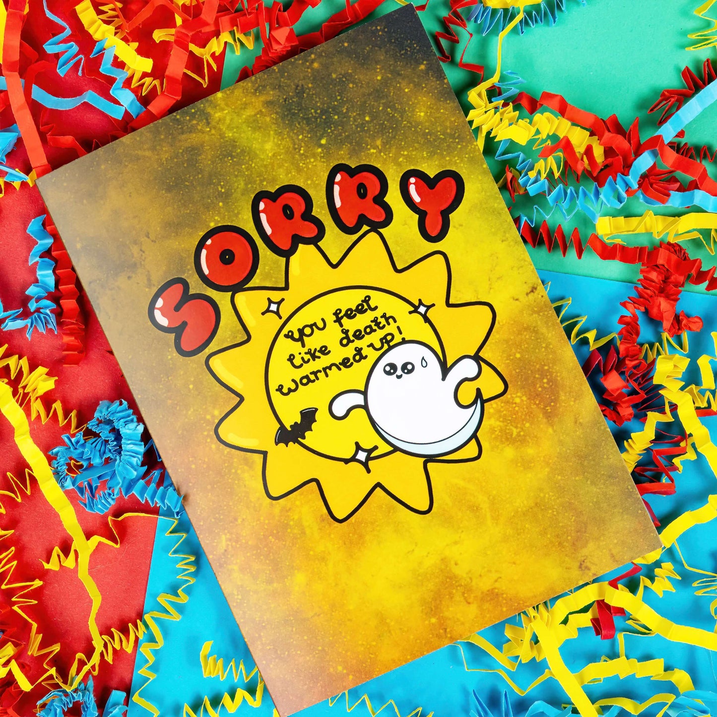 A yellow greeting card with black paint splatters. 'Sorry' is written in big red letters above an illustration of a sun with 'You feel like death warmed up!' inside. there are also sparkles, a sweating ghost and a bat around the sun. The background of the photo is colourful card confetti. The spooky design is inspired by get well soon cards.