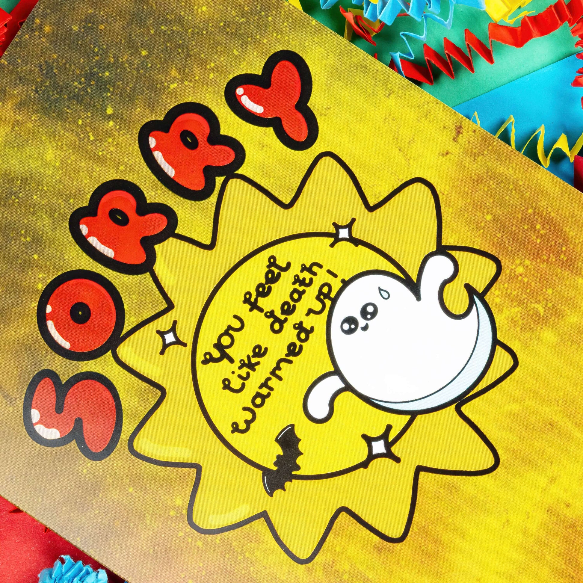 Close up of A yellow greeting card with black paint splatters. 'Sorry' is written in big red letters above an illustration of a sun with 'You feel like death warmed up!' inside. there are also sparkles, a sweating ghost and a bat around the sun. The background of the photo is colourful card confetti. The spooky design is inspired by get well soon cards.