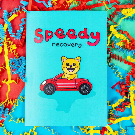 The Speedy Recovery Get Well Soon Cat Card on a colourful card confetti background. The blue card has 'speedy' written at the top in big red letters with 'recovery' written in a smaller black font underneath. In the centre of the card is an adorable orange cat with an open mouthed smile to reveal it's pink tongue. The cat has pink ears and nose and big eyes. The cat is driving a red car.