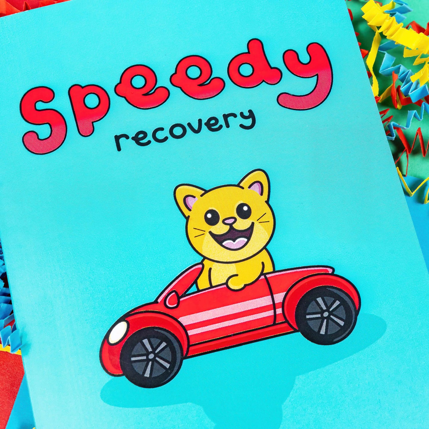 The Speedy Recovery Get Well Soon Cat Card on a colourful card confetti background. The blue card has 'speedy' written at the top in big red letters with 'recovery' written in a smaller black font underneath. In the centre of the card is an adorable orange cat with an open mouthed smile to reveal it's pink tongue. The cat has pink ears and nose and big eyes. The cat is driving a red car.