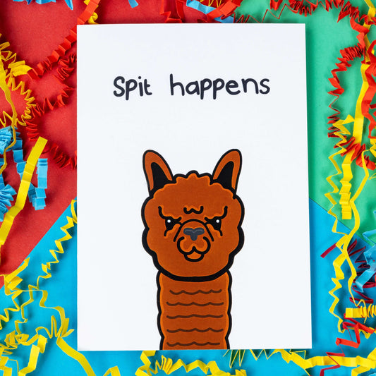 The Spit Happens Alpaca Card on a red, blue and green background red, yellow and blue crinkle card confetti. The white a6 greeting card has an angry brown alpaca on the front with top black text reading 'spit happens'.