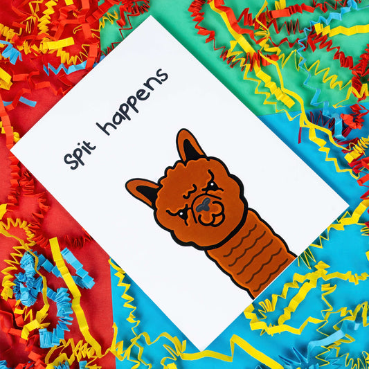 The Spit Happens Alpaca Card on a red, blue and green background red, yellow and blue crinkle card confetti. The white a6 greeting card has an angry brown alpaca on the front with top black text reading 'spit happens'.