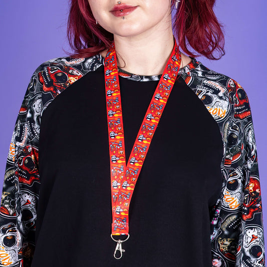 model with red hair and lip piercing wearing a raglan black top with a red lanyard with Innabox Spoonie characters, spoons, rainbows and lighting bolts. She is standing by a purple background