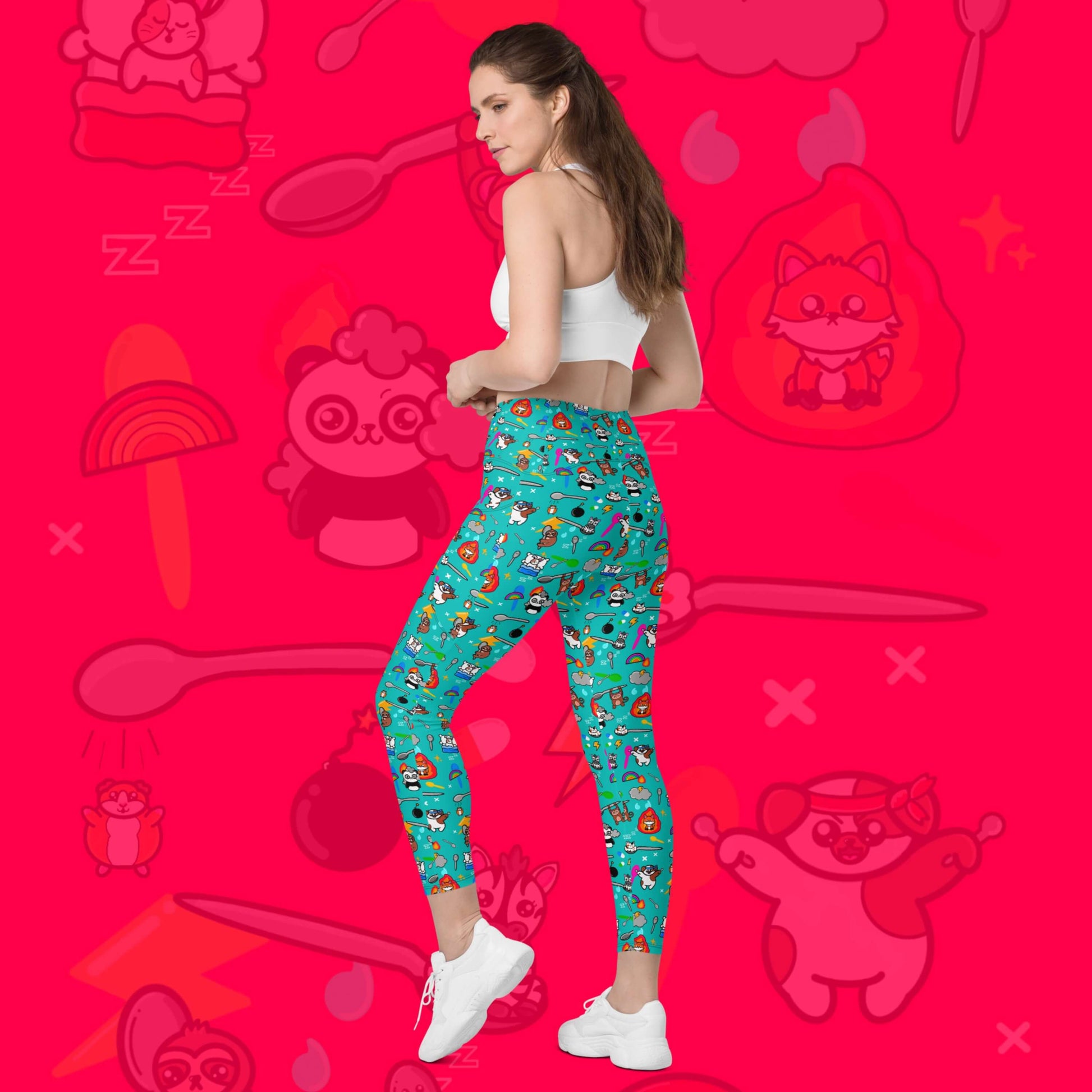 The Spoonie Leggings with Pockets modelled by a femme person with brown hair, white crop top and white trainers on a red background with a faded version of the print in the background. The blue leggings feature various invisible illness themed animals, rainbows, spoons, sparkles, flames, pills and lightning bolts. Raising awareness for hidden disabilities.