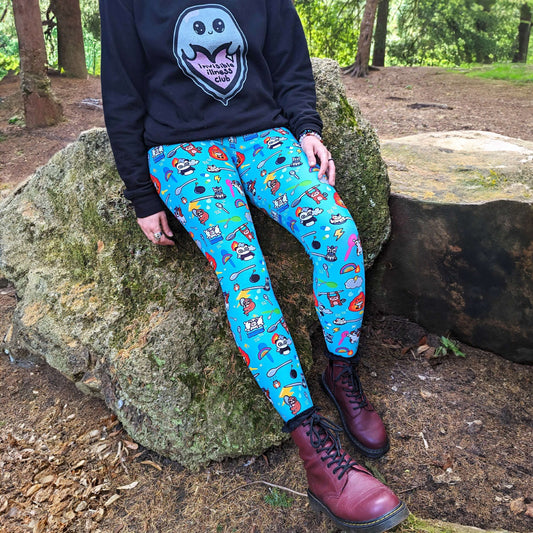 The Spoonie Leggings modelled by Nikky outside in a forest area. She is leaning back on a rock wearing the spoonie leggings with red dr martens and black invisible illness club ghostie jumper. The blue leggings feature various invisible illness themed animals, rainbows, spoons, sparkles, flames, pills and lightning bolts. Raising awareness for hidden disabilities. 
