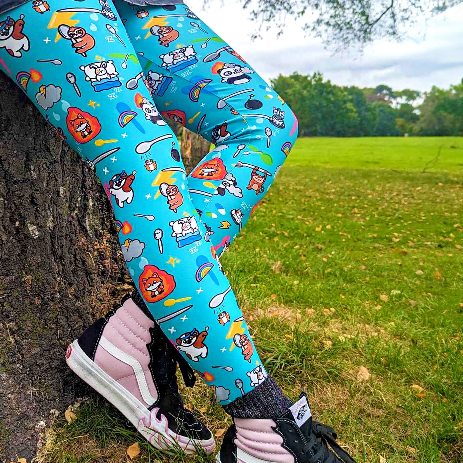 The Spoonie Leggings modelled by Nikky outside in a forest area. She is leaning back on a tree stump wearing the spoonie leggings with pin and black vans trainers. The blue leggings feature various invisible illness themed animals, rainbows, spoons, sparkles, flames, pills and lightning bolts. Raising awareness for hidden disabilities.