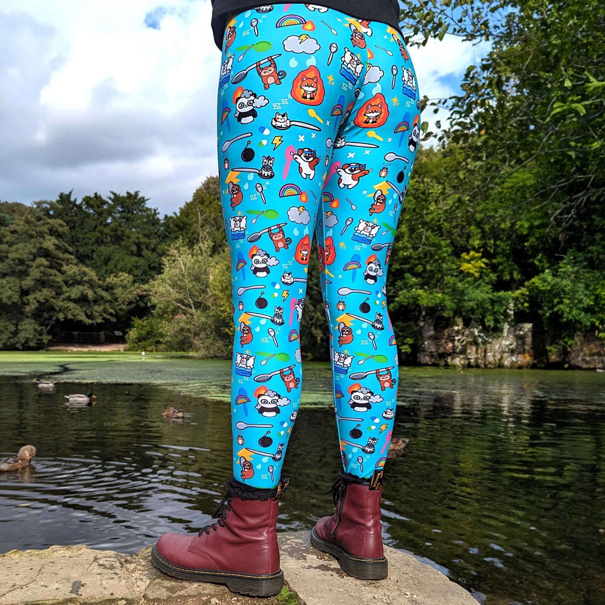 The Spoonie Leggings modelled by Nikky outside in a forest area. She is standing on a rock in front of a lake wearing the spoonie leggings with red dr martens and black invisible illness club ghostie jumper. The blue leggings feature various invisible illness themed animals, rainbows, spoons, sparkles, flames, pills and lightning bolts. Raising awareness for hidden disabilities.