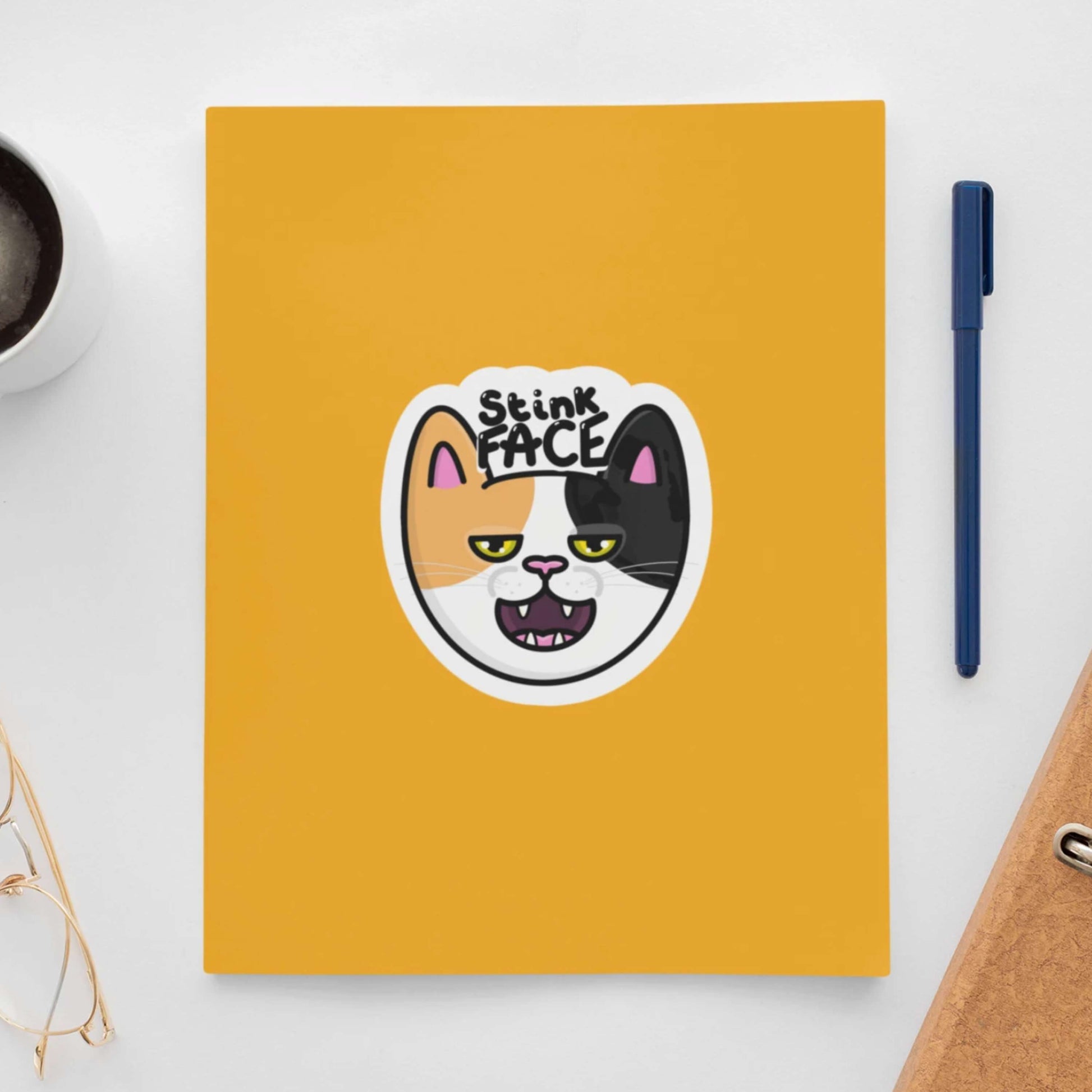 The Stink Face Cat Sticker stuck on a yellow notebook with a black coffee, glasses, file and a pen all on a white desk. The orange and black cat head sticker is smiling with a smug look on its face and black text above reading 'stink face'.