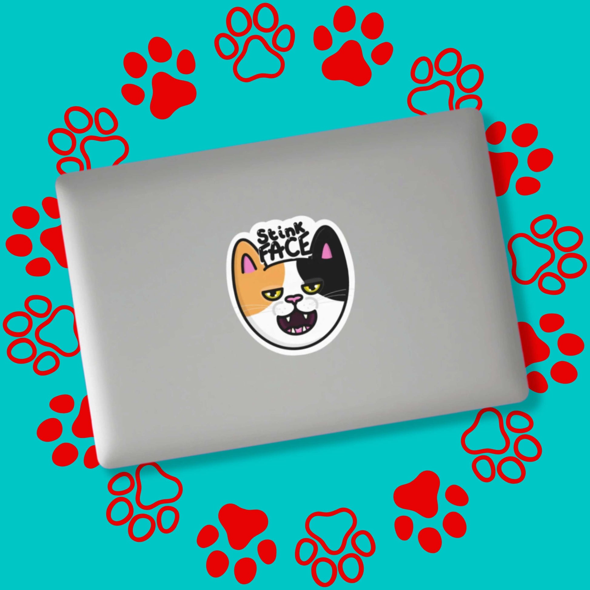 The Stink Face Cat Sticker stuck on a silver laptop on a red and blue paw print background. The orange and black cat head sticker is smiling with a smug look on its face and black text above reading 'stink face'.