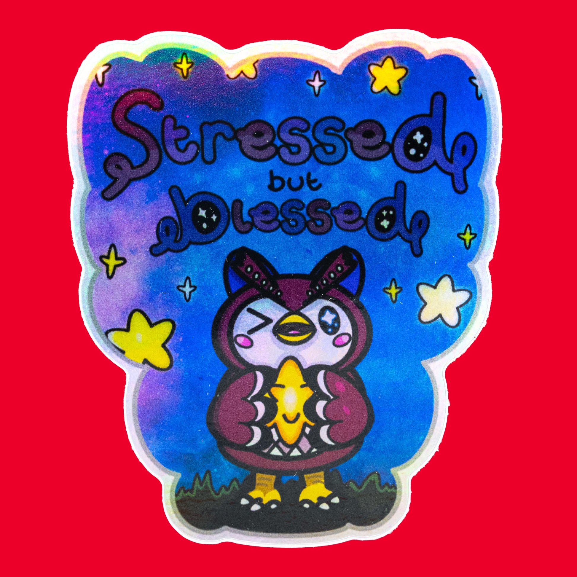 A holographic vinyl sticker by Innabox featuring a cute owl-like creature holding a glowing star with the phrase Stressed But Blessed above it. The colourful galaxy background shines with holographic effects and soft yellow stars. The sticker has a custom cut shape and uplifting disabled and chronic illness inspired design.