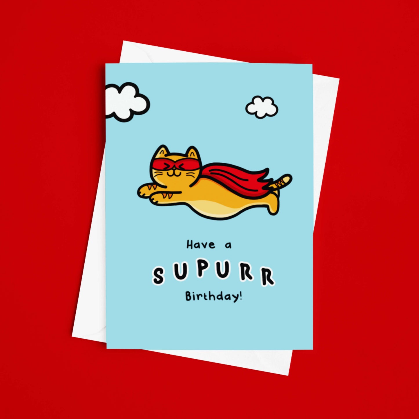 The Have A Supurr Birthday Cat Card on a red background with a white envelope underneath. The pastel blue a6 birthday card of a smiling orange cat flying through a cloudy sky wearing a superhero cape and mask, underneath in black text reads 'have a supurr birthday!'.
