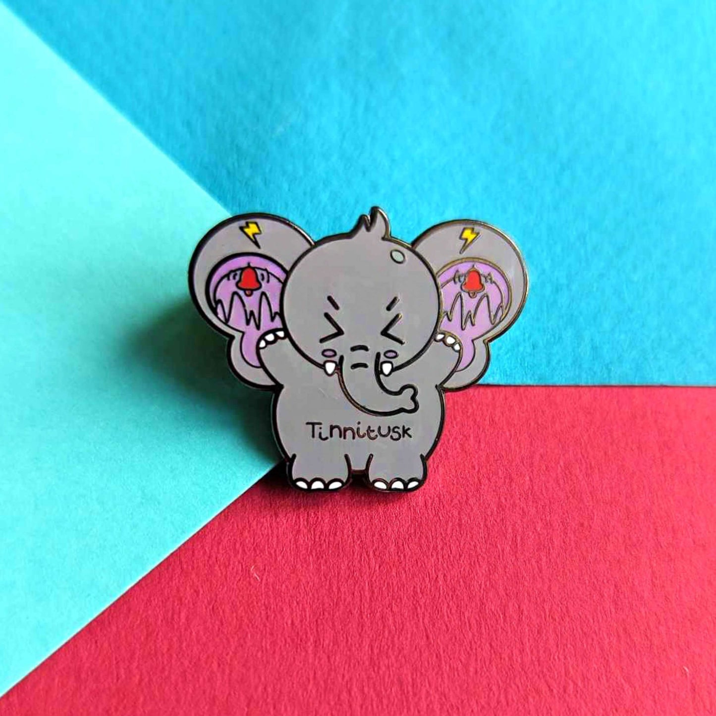 Tinnitus Enamel Pin on a blue and red background. A grey elephant shaped enamel pin with big ears with purple on the inside of them and black squiggly lines with red ringing alarm bells above the lines and yellow lightening bolts above the bells. The elephant has its eyes screwed shut and its arms up. 'Tinnitusk' is written in black across its middle.
