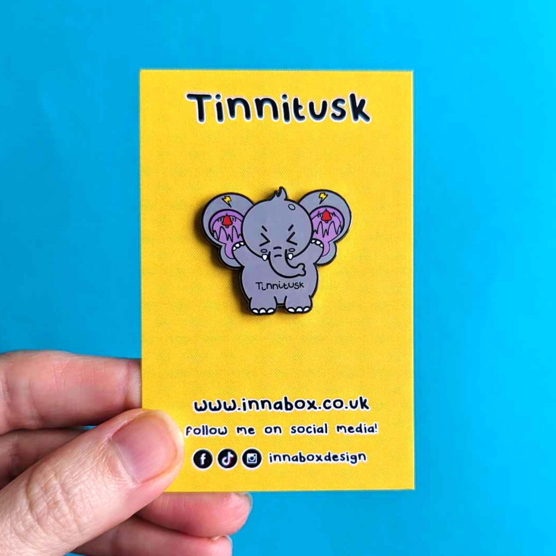 Tinnitus Enamel Pin on yellow backing card being held over a blue background. A grey elephant shaped enamel pin with big ears with purple on the inside of them and black squiggly lines with red ringing alarm bells above the lines and yellow lightening bolts above the bells. The elephant has its eyes screwed shut and its arms up. 'Tinnitusk' is written in black across its middle.